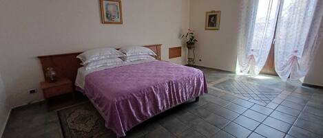Um quarto com cama, mesa de cabeceira, mesinha, cadeira, tapete e janela com cortinas.