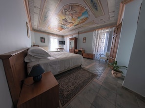 A bedroom with a large bed, a wooden nightstand, a desk, a chair, a television, and a ceiling mural.