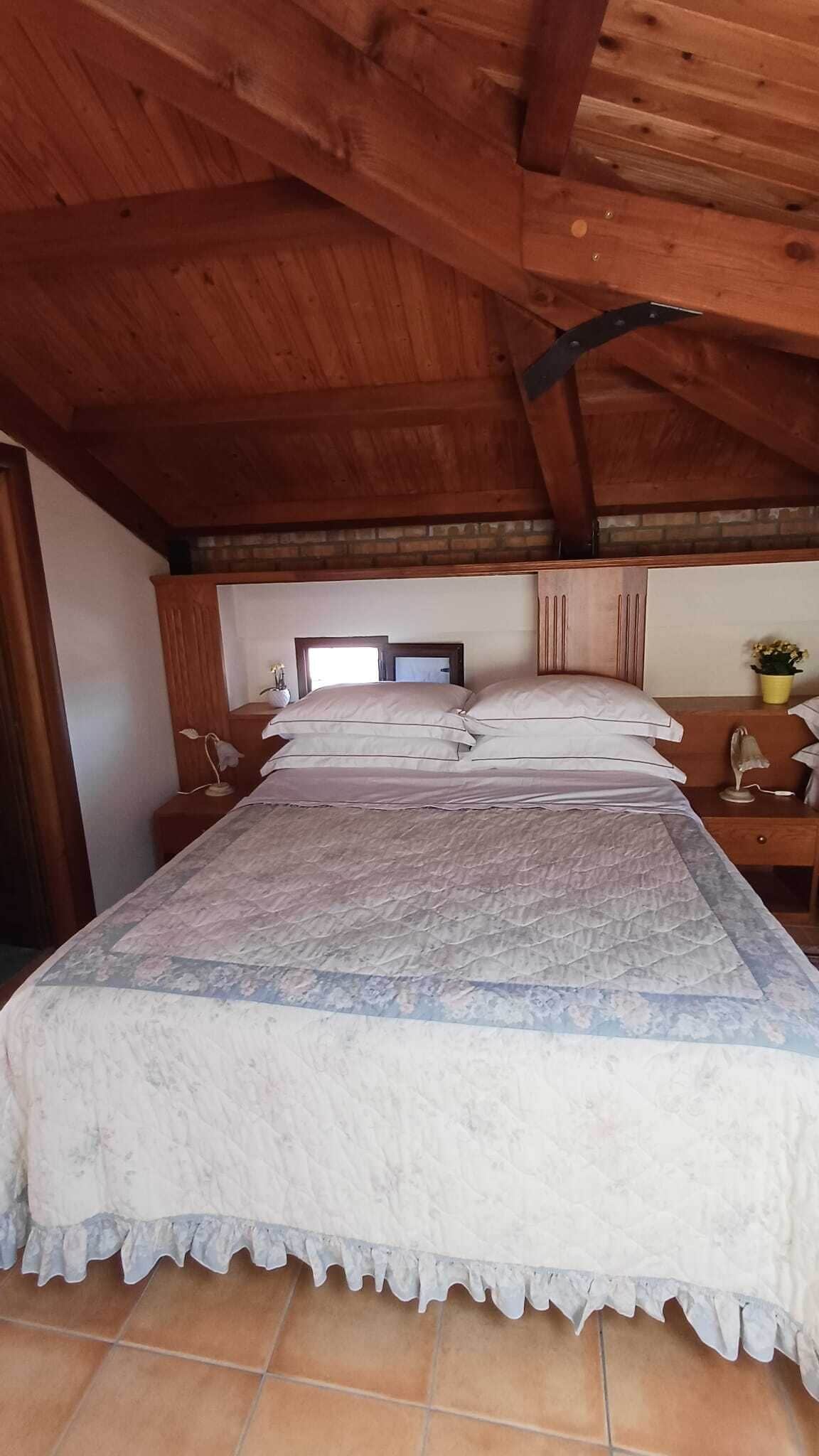 Une chambre avec un plafond en bois, un grand lit recouvert d’une couvre-lit à motifs floraux, une table de chevet avec une lampe et une petite fenêtre.