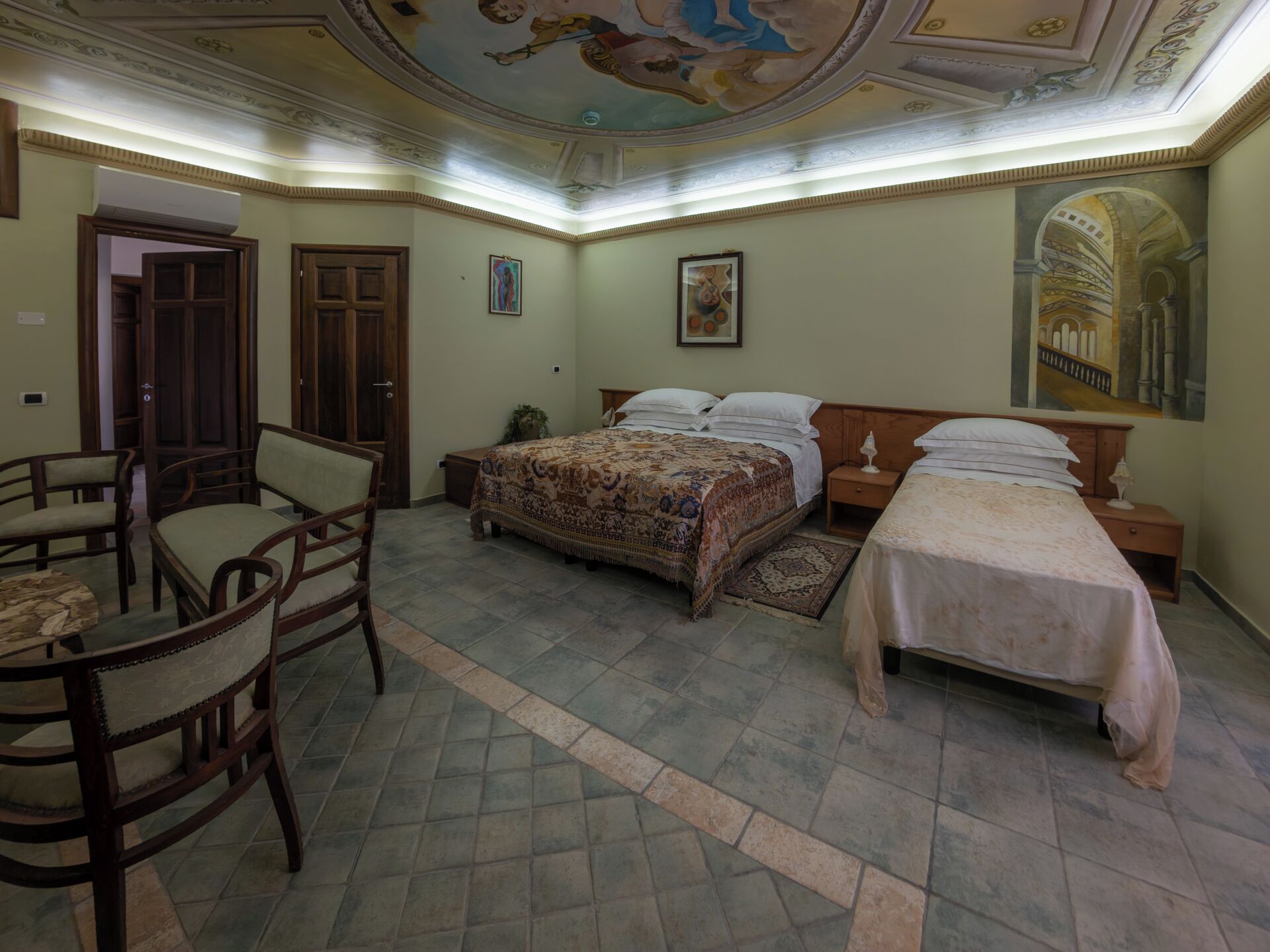 A bedroom with two beds, a chair, a table, and a ceiling mural.