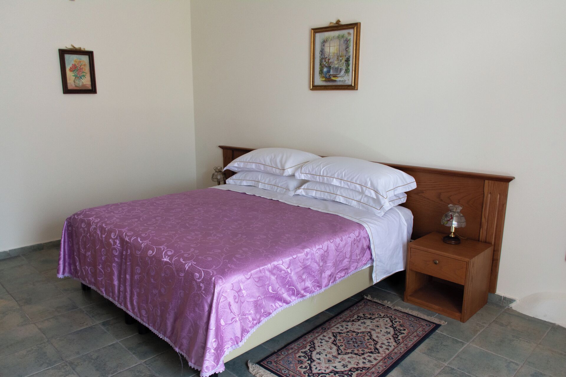 A neatly made bed with a purple bedspread, a wooden nightstand with a lamp, and a patterned rug on a tiled floor.