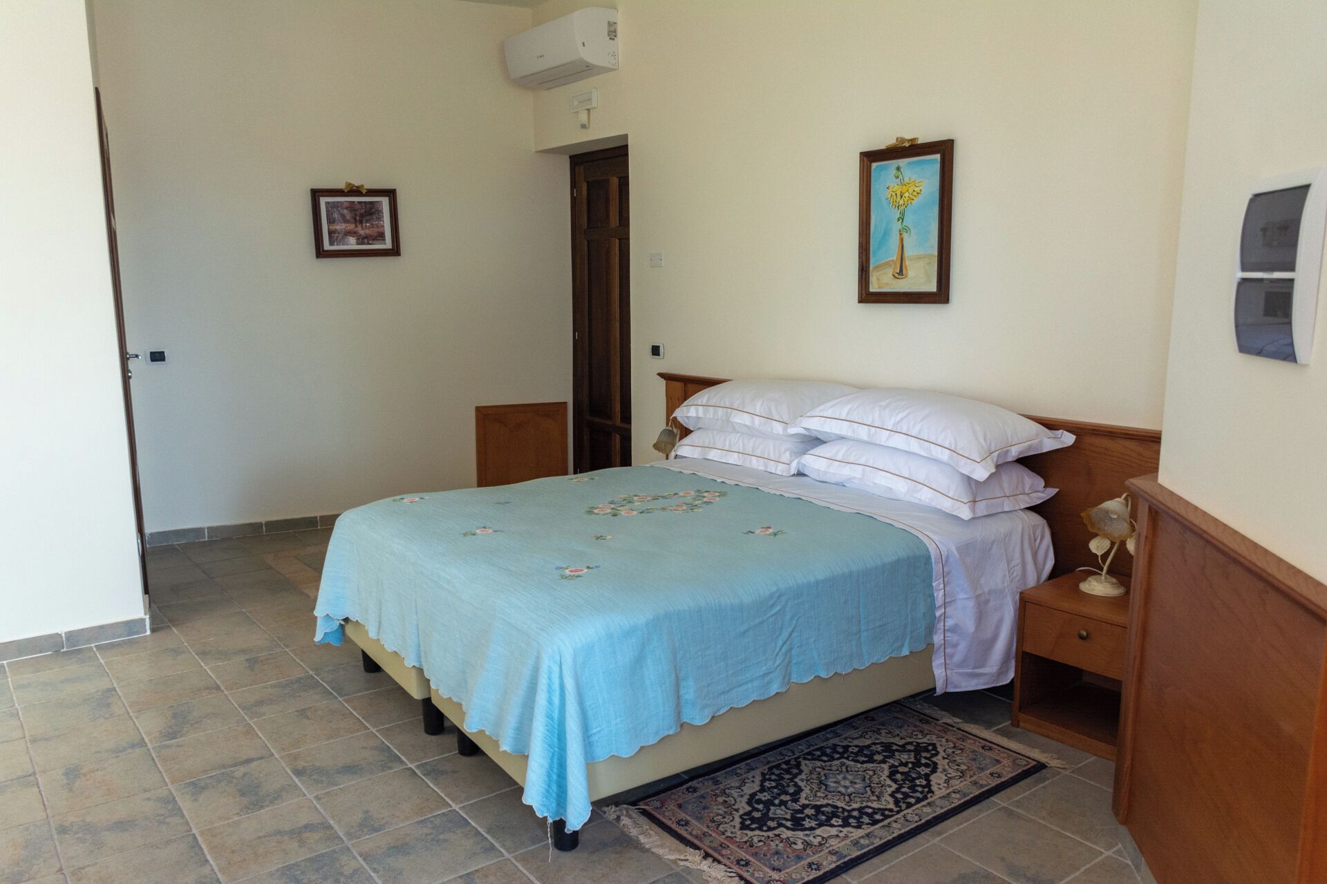 Een slaapkamer met een bed, nachtkastje, een wandairconditioner en een deur naar een andere kamer.