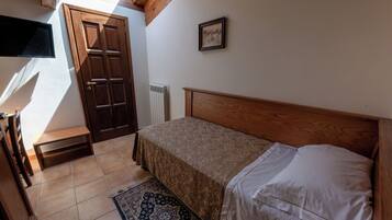 A bedroom with a wooden ceiling, a bed with white linens, a small table, a chair, a radiator, and a window.