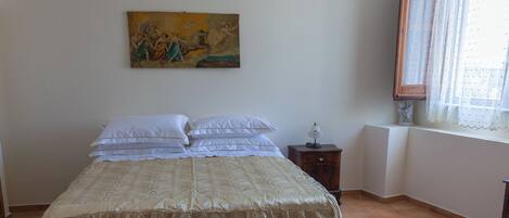 A bedroom with a bed, pillows, a nightstand, a lamp, a painting, and a window with curtains.