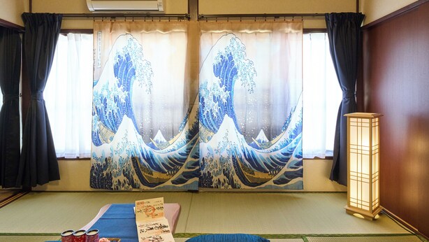 Een traditionele Japanse kamer met tatami vloerbedekking, shoji schermen met golfschilderingen, een tatami mat in het midden, een staande lamp en een kleine tafel met spullen.
