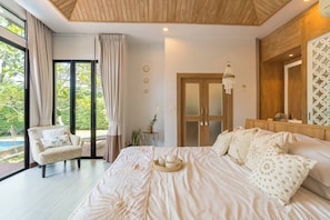Ein Schlafzimmer mit einem großen Bett, einem Sessel, einer Holzbalkendecke, einer Tür und Blick ins Grüne.
