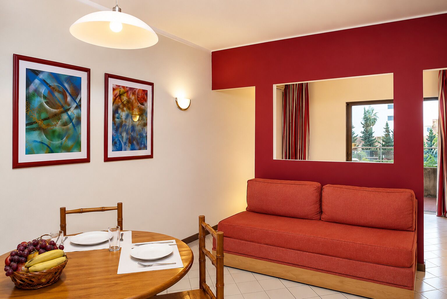 Un comedor con una mesa de madera dispuesta para dos personas, un sofá rojo y dos cuadros abstractos en la pared.