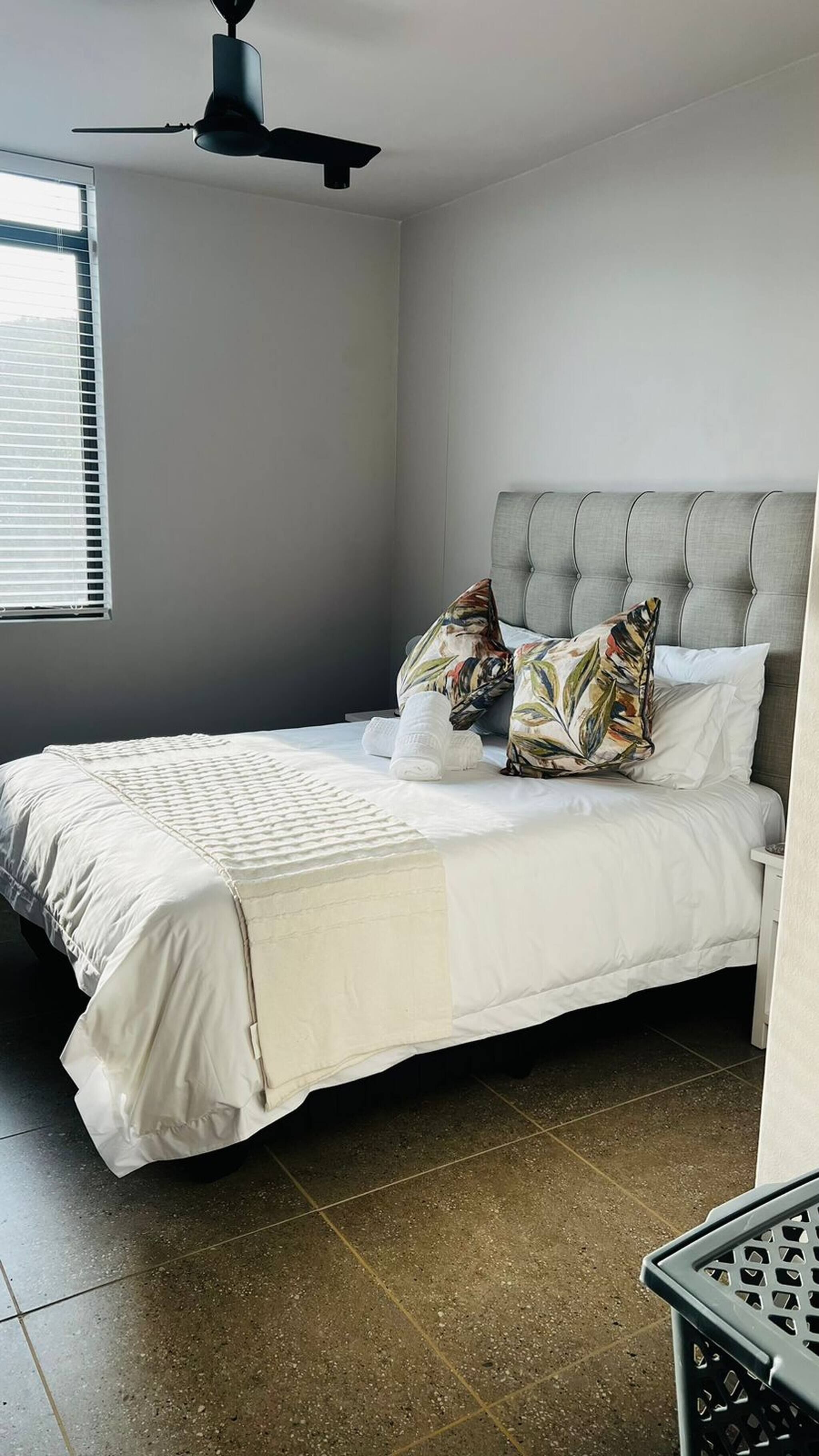 Un dormitorio con cama, almohadas, ventilador de techo y ventana.