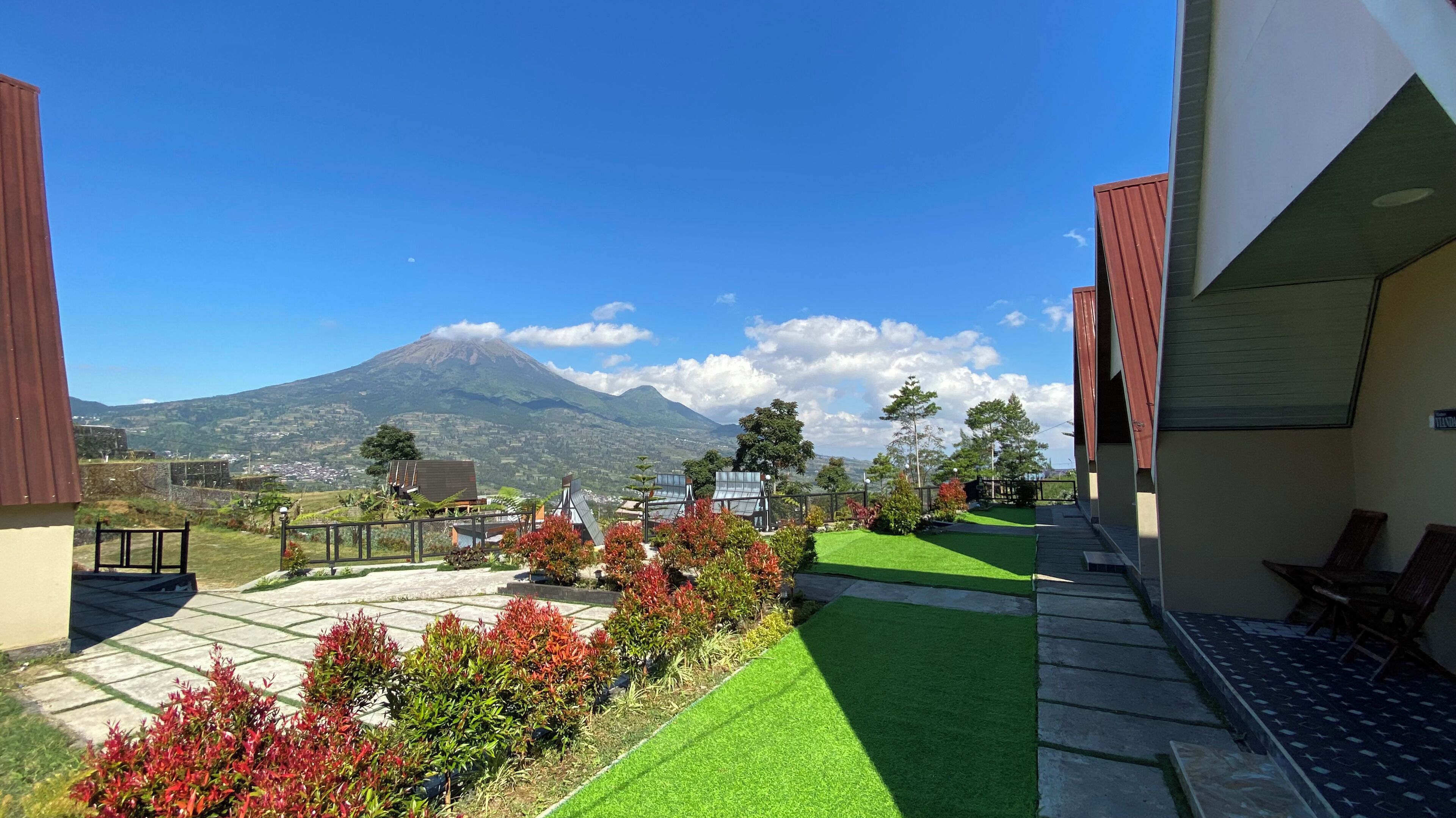 Vista de una montaña con cielo despejado, un jardín cuidado con césped artificial y plantas coloridas, y un edificio con techo inclinado.