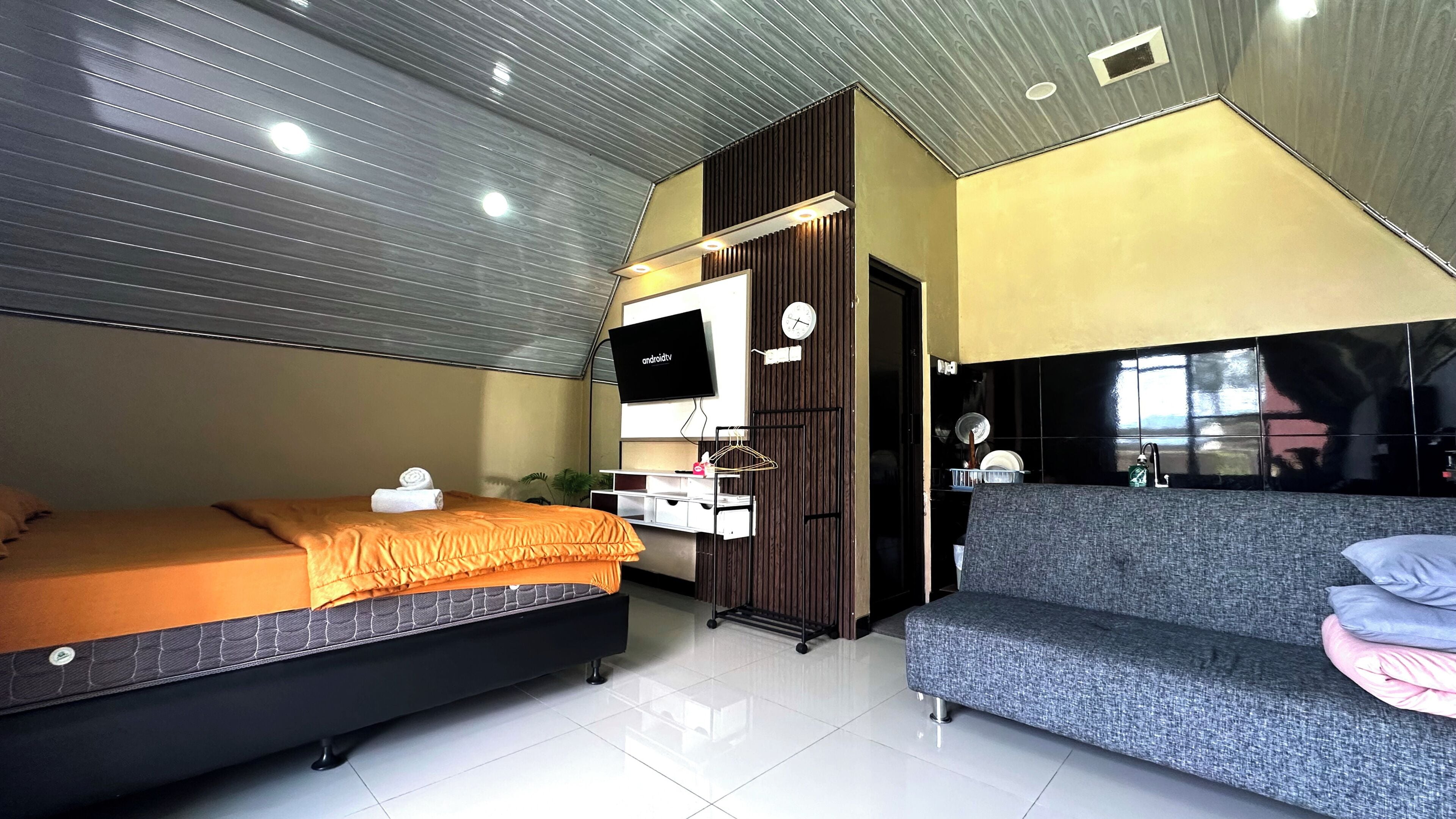 A modern hotel room with a bed, a sofa, a TV, a small kitchenette, and a window.