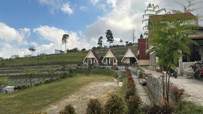 Un entorno rural con un camino que lleva a una hilera de pequeños edificios de dos pisos con techo inclinado, rodeado de follaje y un cielo despejado.