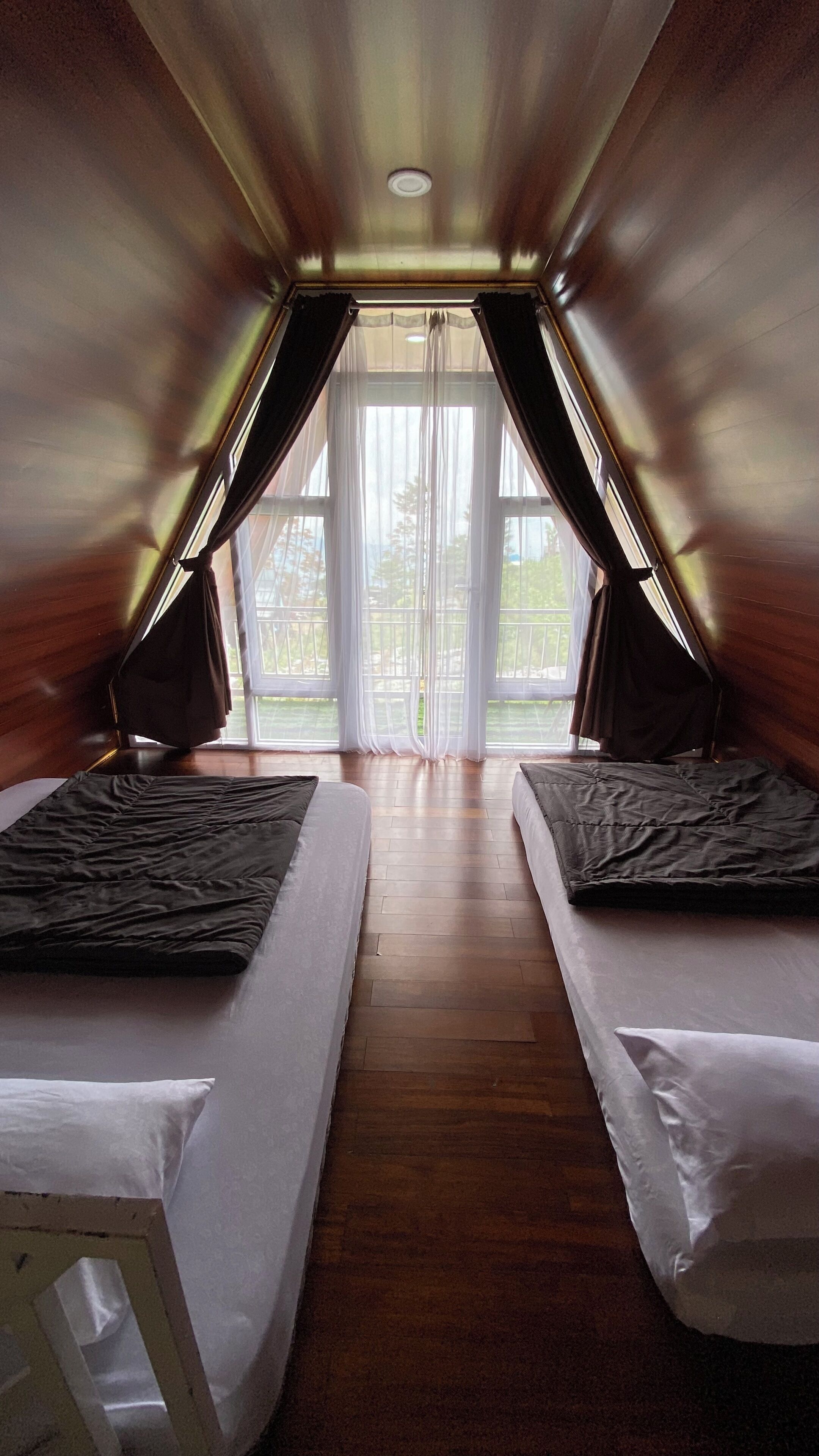 A room with two beds, a wooden ceiling, and a large window with curtains.