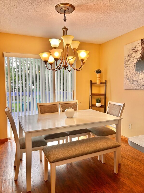 A dining area with a table, chairs, a bench, a chandelier, and a view through a window with blinds.
