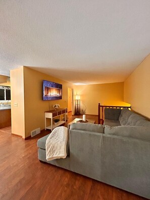 Ein Wohnzimmer mit einer Couch, einem an der Wand montierten Fernseher, einer Lampe und einem kleinen Tisch mit einer Pflanze.