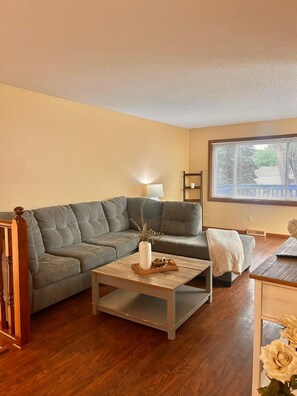 Ein Wohnzimmer mit grauem Sofa, einem Holztisch mit Vase, einer Lampe und einem Fenster mit Jalousien.