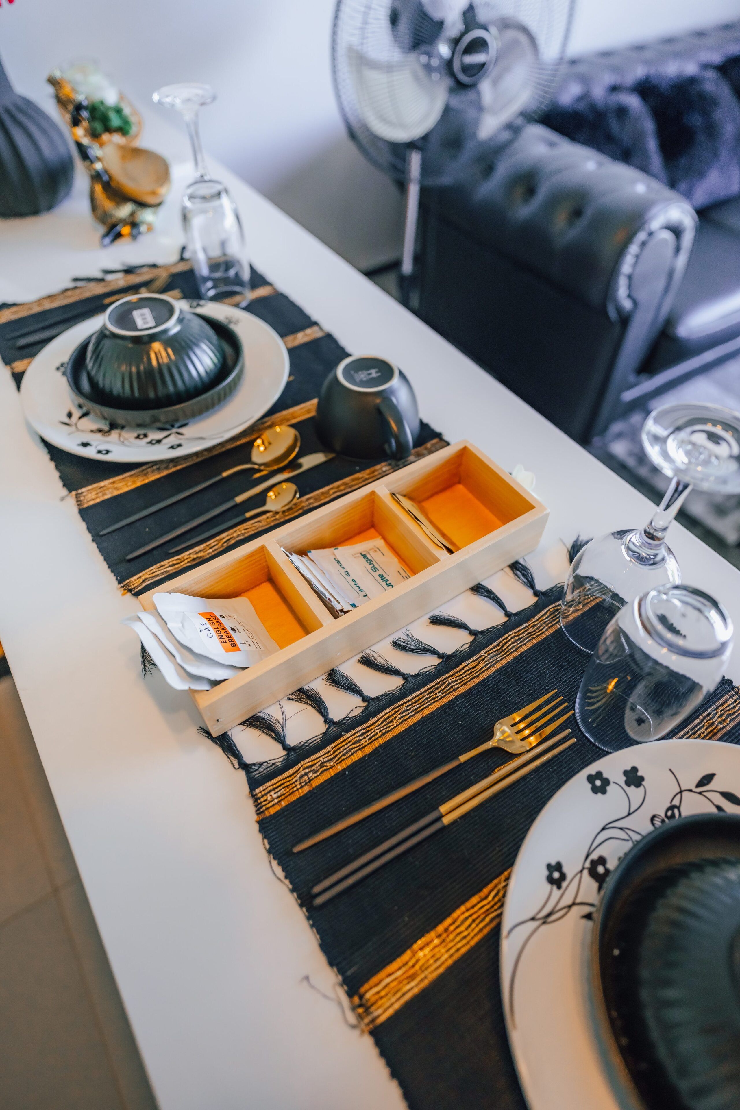 A dining table with a placemat, plates, cutlery, and a fan in the background.