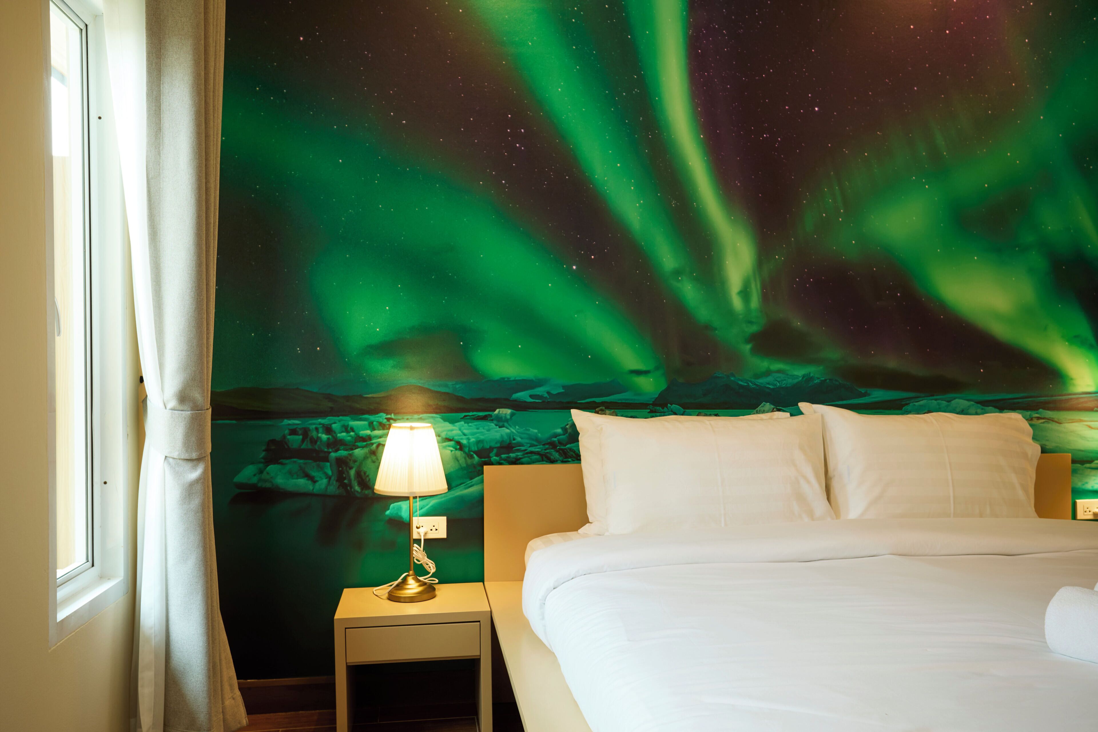 A hotel room with a bed, a nightstand with a lamp, and a wall mural of the Northern Lights.