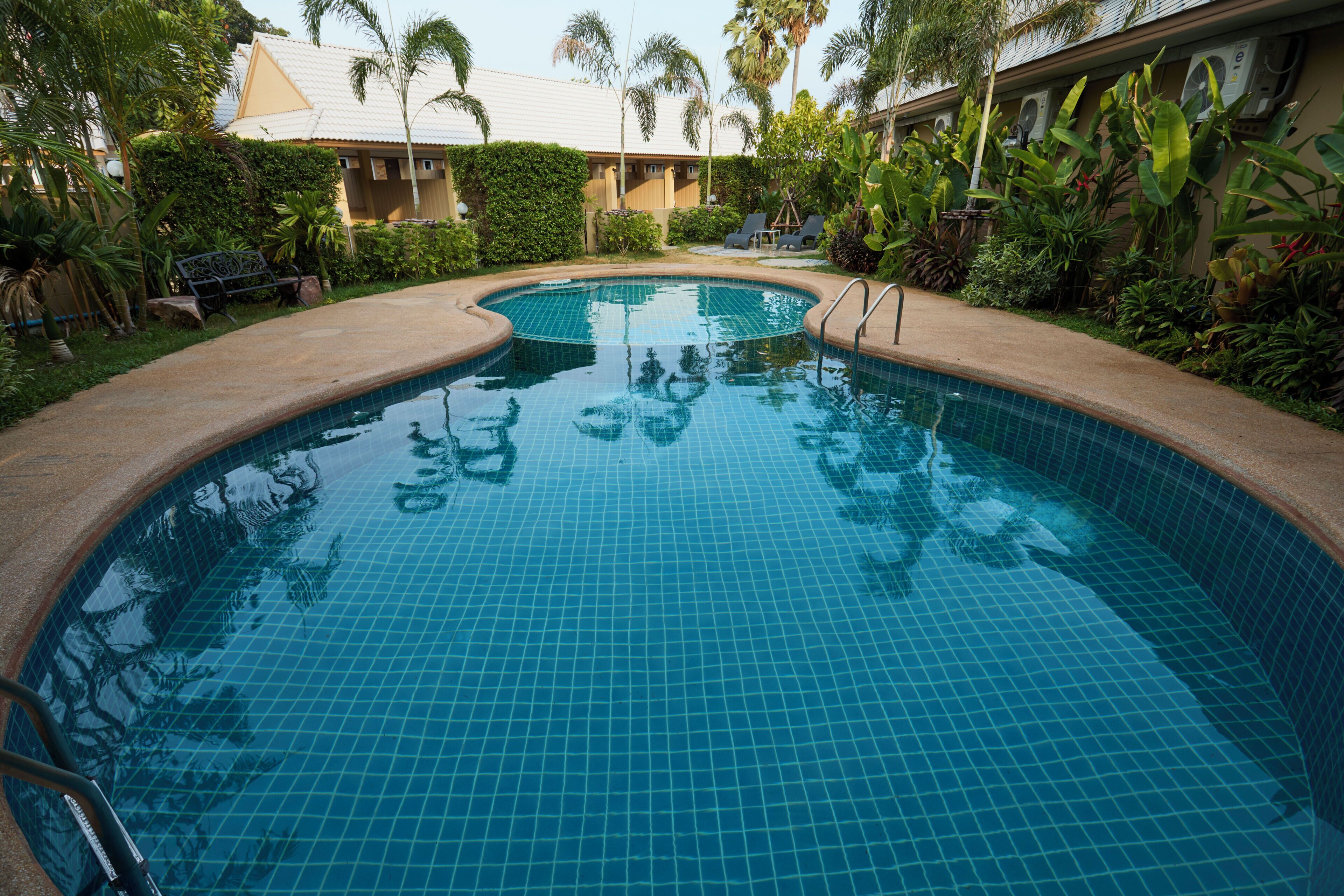 Una piscina con superficie de azulejos, rodeada de vegetación y un edificio con techo de tejas.