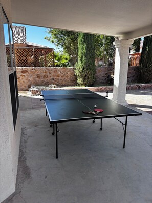 Ein Tischtennistisch im Freien mit Netz, Ball und Schlägern vor einer Steinwand und Grünpflanzen.
