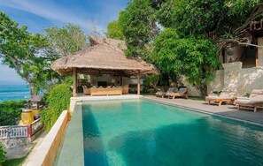 Ein Pool mit Liegestühlen, einer Struktur mit Reetdach und Blick aufs Meer.