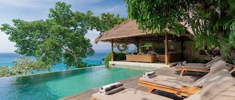 A wooden deck with lounge chairs and a thatched-roof structure overlooking a pool.