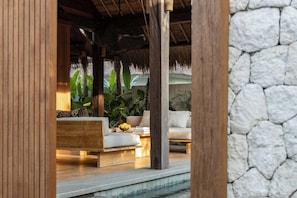 A wooden-framed room with a stone wall, a thatched roof, and a seating area with cushions and a table. - Villa Kokomo - 2 Bedroom Villa (Nusa Lembongan)