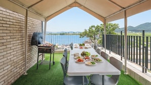 Un área de comedor al aire libre con una parrilla, una mesa dispuesta con distintos platos y vistas al agua y colinas.