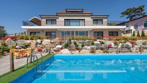 Una casa moderna de dos pisos con piscina, mobiliario exterior y un jardín ajardinado con rocas y flores.