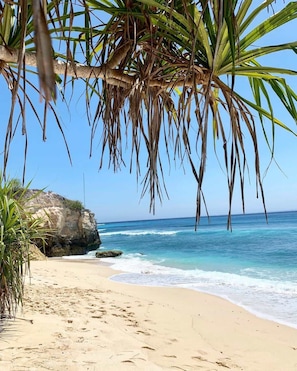 Ein Strand mit klarem, blauem Wasser, sandigem Ufer und einer großen Felsformation.
