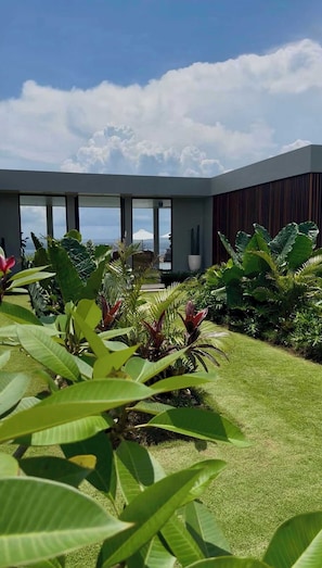 Ein modernes Gebäude mit großen Fenstern, ein gepflegter Garten mit tropischen Pflanzen und Blick aufs Meer.