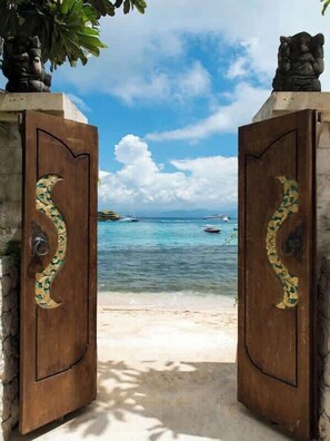 Ein hölzernes Tor mit Zierde, das zu einem Strand mit klarem Wasser und entfernten Booten führt.