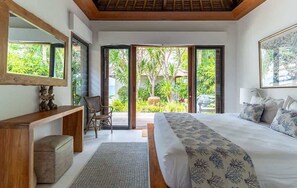 Ein Schlafzimmer mit einem großen Bett, einem Holztischchen, einem Sessel und Blick auf Grün hinter einer Glasdoor.