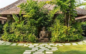 Eine Statue einer sitzenden Figur, umgeben von üppiger Vegetation und einem Gebäude mit einem Reetdach.