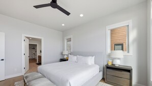 Une chambre à coucher avec un lit, des tables de chevet, un banc, un ventilateur de plafond et une fenêtre.