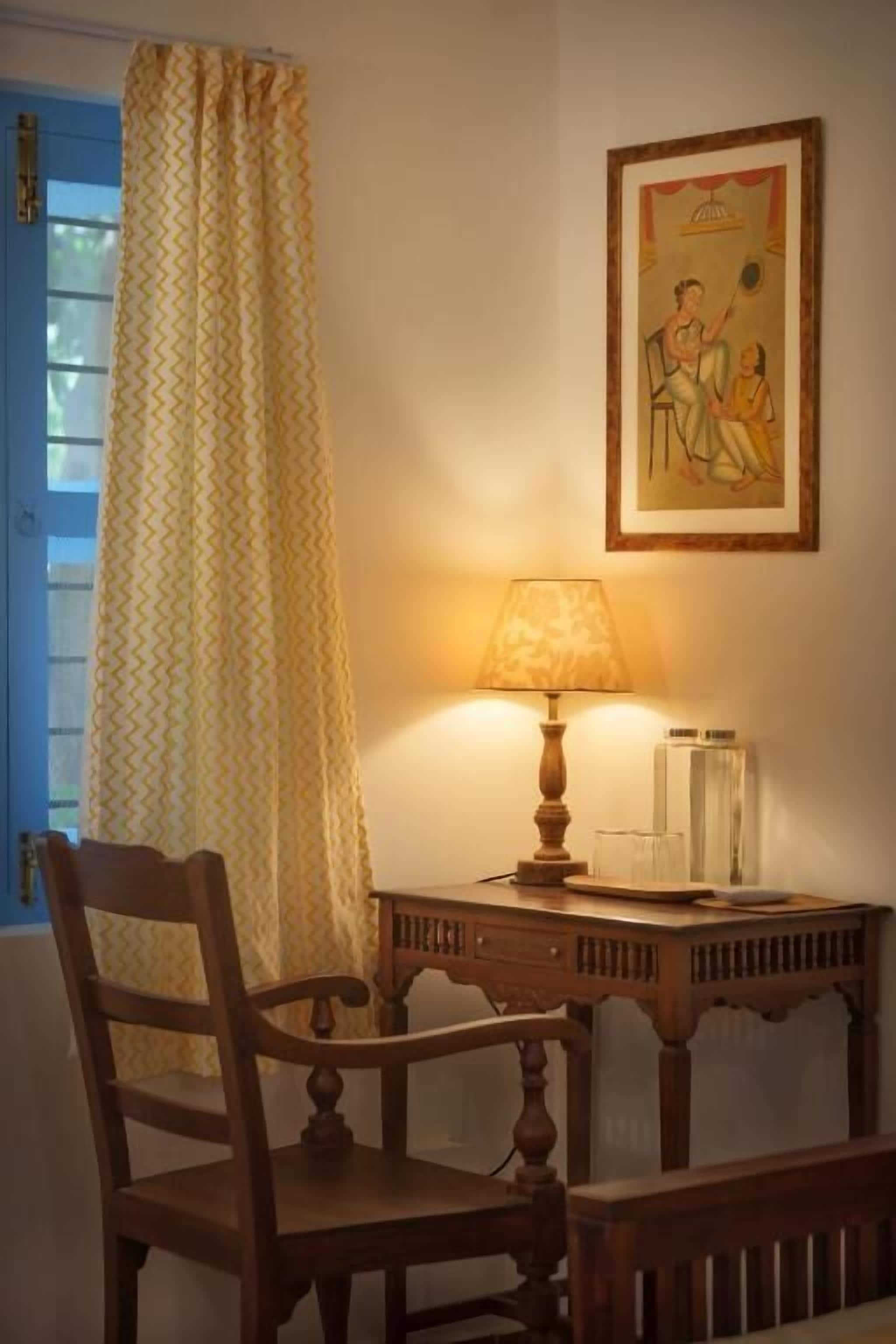 A room with a wooden chair, a table with a lamp, a framed picture on the wall, and a window with curtains.