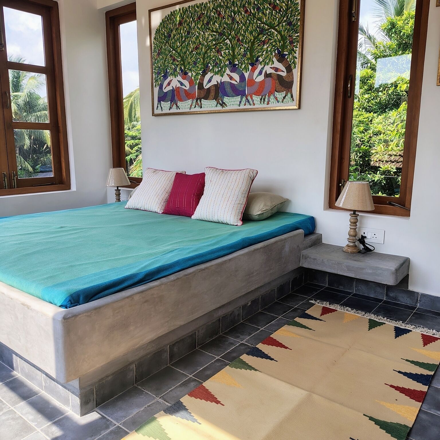 Une chambre à coucher avec un lit, des oreillers, une table de chevet, une lampe, un tableau et une vue sur la verdure.