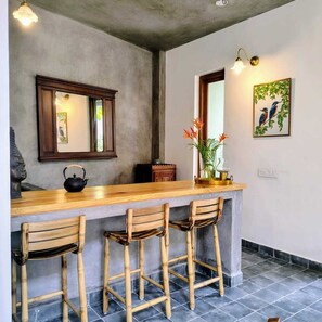 A kitchen with a wooden bar, three bamboo chairs, a black kettle, and a vase with flowers. - Isai Karai Bungalow - A CGH Earth Saha (Tirupporur)