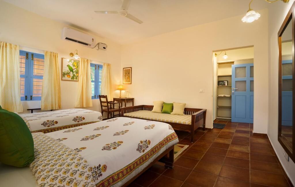 Une chambre avec deux lits, un ventilateur de plafond, une fenêtre avec des rideaux, un bureau avec une chaise et une petite table avec une lampe.