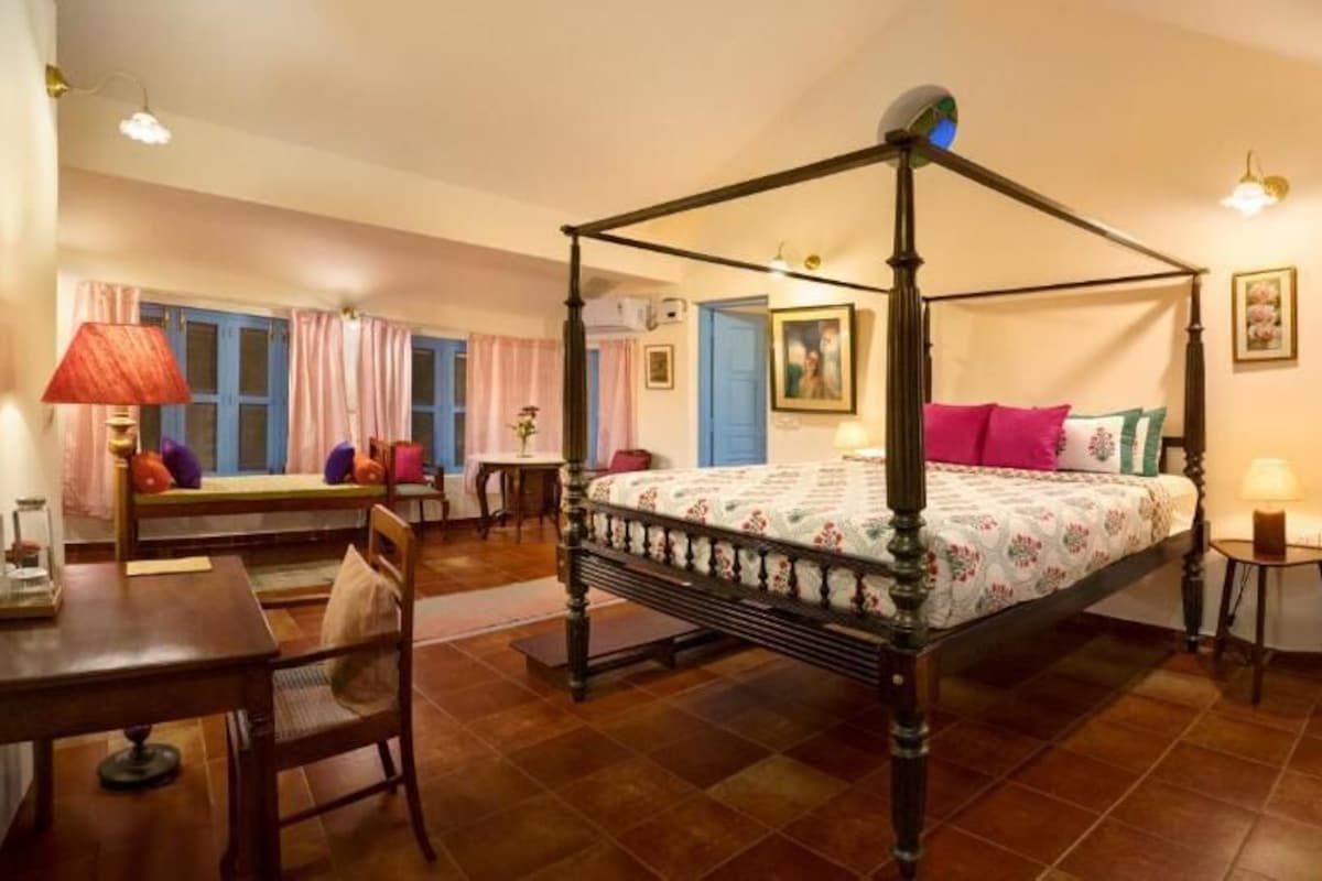 Una cama con dosel en una habitación con suelo de madera, una mesa de comedor, un sofá y una mesita de noche con una lámpara.