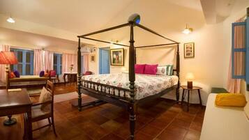 A four-poster bed in a room with a wooden floor, a dining table, a sofa, and a small table with a lamp.