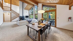 A spacious living area with a wooden ceiling, a dining table set for four, and a large window overlooking a deck. - Forest Views + Wrap Decks | Truckee + Lake Tahoe | Bear's Lair (Truckee)