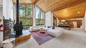 A spacious living area with a fireplace, large windows, a kitchen, and a dining area. - Forest Views + Wrap Decks | Truckee + Lake Tahoe | Bear's Lair (Truckee)