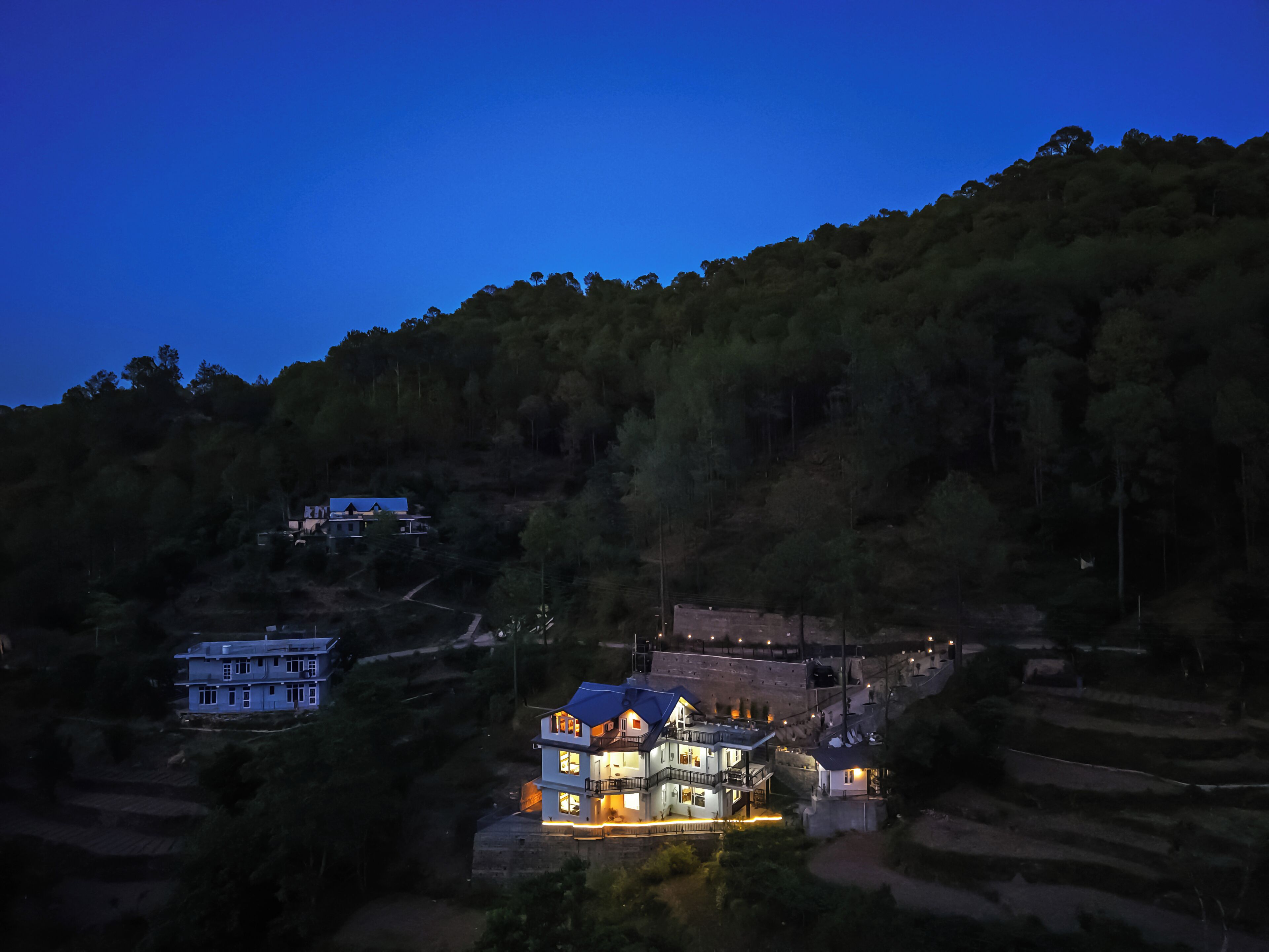 A large, multi-story house with a prominent balcony, surrounded by terraced fields and dense forest.