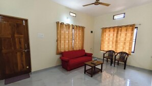 A room with a wooden door, a red sofa, two wooden chairs, a small table, and a ceiling fan. - Anthik Family Homestey (Bhatkal)