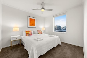 A hotel room with a bed, two bedside tables, a ceiling fan, a window with a city view, and a framed picture on the wall. - Exclusive Stays - Rivergarden (Southbank)
