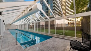 Una piscina interior con techo de cristal, desde donde se ve una cancha de tenis, y una silla negra.