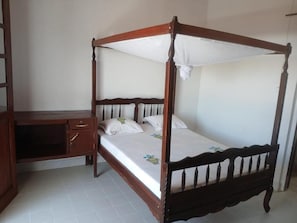 Una cama con dosel sobre cuatro postes, un escritorio de madera con cajones y un suelo de baldosas blancas.