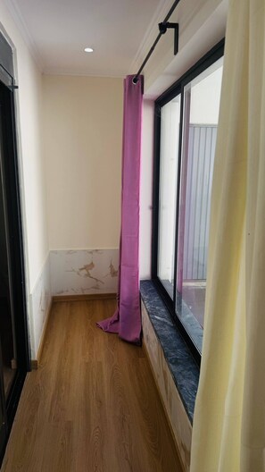 Een smalle gang met een houten vloer, een raam met een zwarte omlijsting en een paars gordijn.