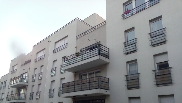 Ein mehrgeschossiges Wohngebäude mit Balkonen, einem verschlossenen Eingangsbereich und einer Hecke davor.