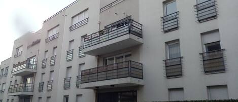A multi-story residential building with balconies, a gated entrance, and a hedge in front.